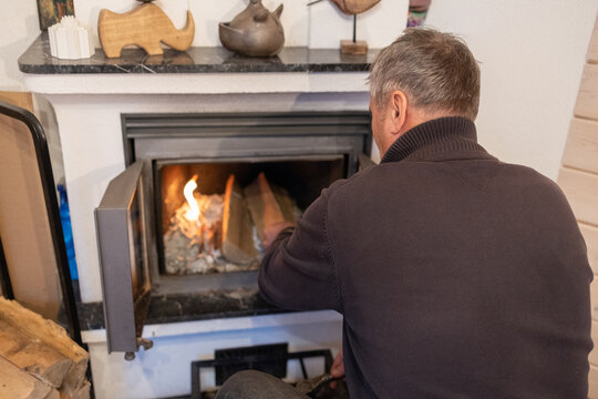 Senior male loading wooden log into stove, lights fireplace inside living. Space winter warmth concept. - Powered by Adobe