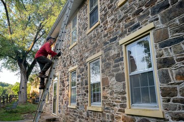 Low angle view of man homeowner climbing dangerous ladder against old stone farmhouse for maintenance cleaning gutters architectural features windows roof 