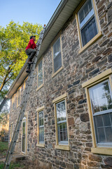 Low angle view of man homeowner climbing dangerous ladder against old stone farmhouse for maintenance cleaning gutters architectural features windows roof 