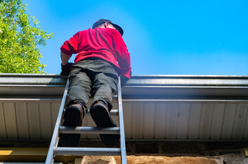Low angle view of man homeowner climbing dangerous ladder against old stone farmhouse for maintenance cleaning gutters architectural features soffit roof 