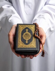 A Man In Traditional White Attire Offers A Detailed Black Quran With Golden Arabic Script And A Tasbih Prayer Beads On Top In A Studio Setting With Soft Lighting