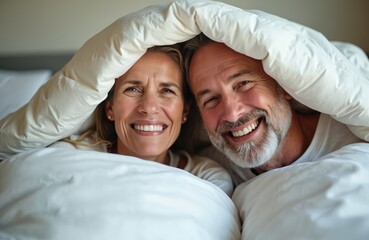 Mature couple laughs under white duvet in bed at home. Happy people share moment of joy and intimacy. Woman and man enjoy cosy morning together, feeling warmth and connection.