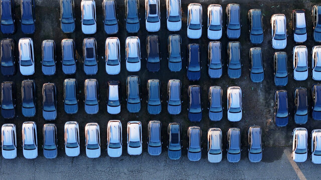 Rows of new cars parked in a large outdoor distribution lot, view from above showing automotive mass production and transportation logistics