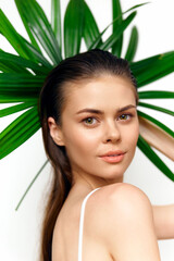 Fototapeta premium Young woman with long hair wearing minimal makeup and white clothing, surrounded by tropical leaves, expressing calm confidence in a bright setting.