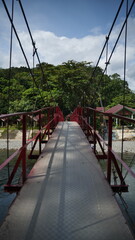 Fototapeta premium Red Metal Suspension Bridge Crossing a River Toward Tropical Village and Forest at Bukit Lawang