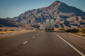 Following behind a silver 18 wheel truck on a two lane highway in the desert with a rocky hill in the distance. The image is from the I-15 interstate in Nevada