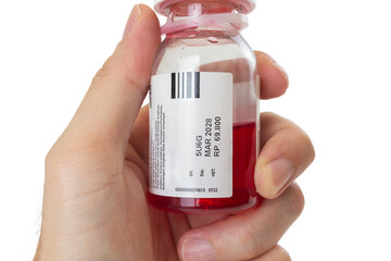 Male hand holding a small transparent plastic bottle of red liquid medicine or syrup. The white label clearly displays the expiration date "MAR 2028", batch number, and price in Indonesian Rupiah.