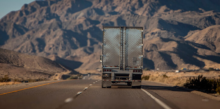 Following behind a silver 18 wheel truck on a two lane highway in the desert with a rocky hill in the distance. The image is from the I-15 interstate in Nevada