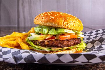 A classic hamburger on a sesame bun with a slice of tomato and lettuce and French fries on the side on lack checked paper