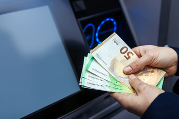 Hands holding various banknotes in front of an ATM machine, showcasing cash withdrawal process with digital display and illuminated buttons in the background