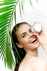 Smiling woman holding skincare cream surrounded by green leaves, showcasing natural beauty and freshness in a tropical setting.