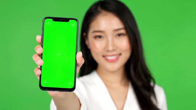 Engaging shot of a cheerful young Asian woman holding up a modern mobile phone with a prominent blank green screen, perfect for creative digital mockups, app demonstrations, or custom content display
