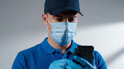 Man in blue uniform, cap, mask, and gloves looks at phone.  Plain background and focus on tech