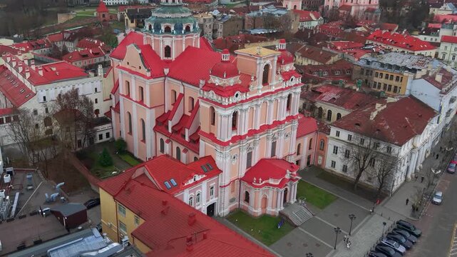 Drone footage captures the stunning Church of St Casimir with vibrant red rooftops amidst the historic architecture and streets of Vilnius Old Town, creating a dynamic aerial perspective.