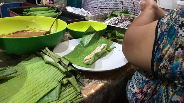 Flavored glutinous sticky rice is rolled and wrapped in a banana leaf to create homemade steamed rice cakes on a cluttered work table by an elderly woman.
