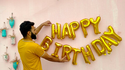 Young bearded man arranging gold foil birthday balloons on a pink wall for party decoration