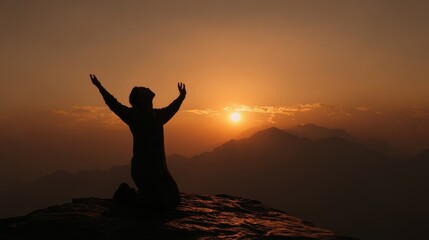 Silhouette of a person kneeling with arms raised towards a vibrant sunset over misty mountains.