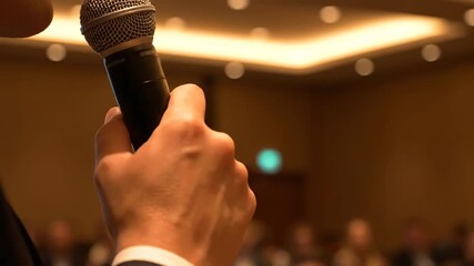 Close up of a hand holding a silver microphone in a conference room with blurred audience under warm lighting, promoting business communication and public speaking.