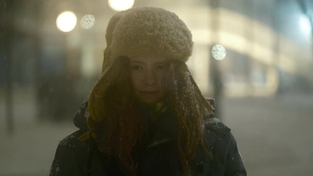 Cinematic night atmosphere with snow falling. A teenage girl with red hair in an ushanka hat looks directly at the camera, creating an intimate, winter portrait