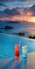 Two vibrant drinks, red and blue, sit by a pool overlooking a tranquil sunset over a tranquil ocean and islands.