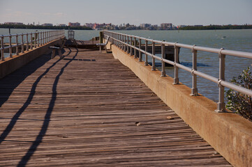 Obraz premium Damaged Long Wooden Pier Over Calm Water With Railings Leading Toward a Distant City Skyline in St. Petersburg, FL Weathered pier extends into tranquil water, with metal railings on both sides. Benche