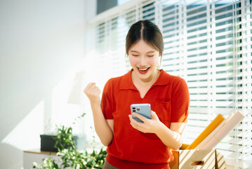 sian woman celebrating personal finance success while checking smartphone at home office. Online business growth, smart money management and positive lifestyle concept.