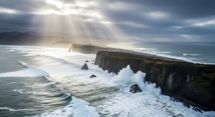 Aerial view of a rugged coastline under a cloudy sky. Sunbeams illuminate the powerful waves crashing against the cliffs