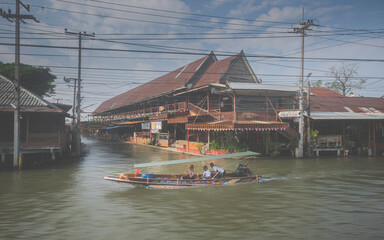 Fototapeta premium A traditional long-tail boat with tourists cruising past a large historic wooden house in a Thai floating market community.