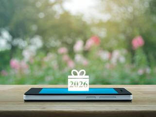 Gift box happy new year 2026 flat icon on modern smart mobile phone screen on wooden table over blur pink flower and tree in park, Business happy new year 2026 shop online concept