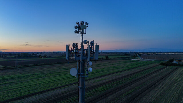 Cell tower providing essential mobile communication technology and data transmission infrastructure across rural Po Valley fields at twilight, connecting remote communities