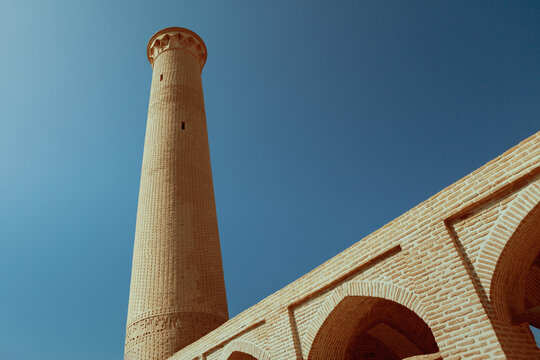 Low-angle view of the Zavareh Minarets in Isfahan, showing a tall ancient brick minaret beside arched structures, highlighting detailed masonry against a clear blue sky.