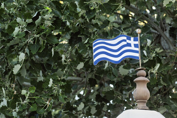 ymbolic scene in Ermoupolis, Syros, Greece, showing a small Greek flag waving gently in front of a leafy green tree. The flag, mounted on a decorative pole, features blue and white stripes 