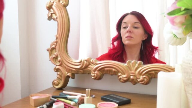 A young woman with vibrant red hair looks contemplatively at her reflection in an ornate golden mirror on a vanity table, surrounded by flowers and makeup, conveying self-reflection and beauty rituals