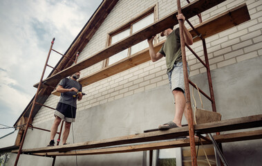 Two men working on scaffolding, installing wooden planks to renovate a house facade. Manual labor, home improvement, and construction project in progress under a cloudy sky.