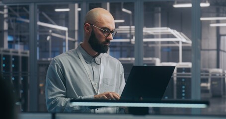 Fototapeta premium Focused IT Engineer Using Laptop in Server Hall. Man in Light Shirt Standing and Working Intently, Background Shows Server Racks Modern Data Center. Concept On Site Diagnosis and System Maintenance.