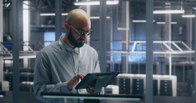 Technician Using Tablet For Asset Management in Data Center. Man Check Assets in Server Hall. Female Colleague is Walking in Background. Concept of Interactive Asset Management and IT Workflow. - Powered by Adobe