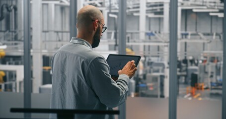 Bearded Male Engineer Analyzing Production Data Graphs on Tablet. Man Standing in Middle of Modern, Automated Manufacturing Facility. Concept of Mobile Data Analysis and Smart Factory Operations.