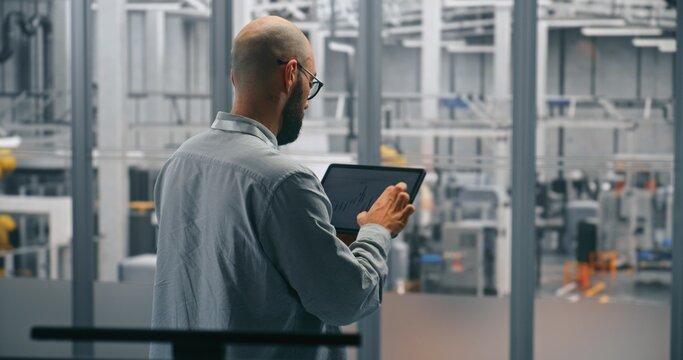 Bearded Male Engineer Analyzing Production Data Graphs on Tablet. Man Standing in Middle of Modern, Automated Manufacturing Facility. Concept of Mobile Data Analysis and Smart Factory Operations. - Powered by Adobe