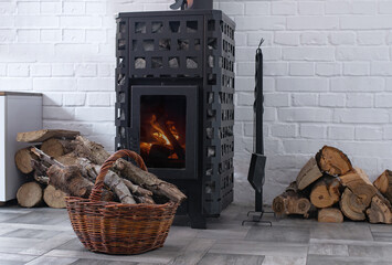 Modern Wood Stove with visible burning fire surrounded by stacked firewood and wicker log basket in a cozy Scandinavian style interior. Home heating, winter comfort and sustainable energy concept