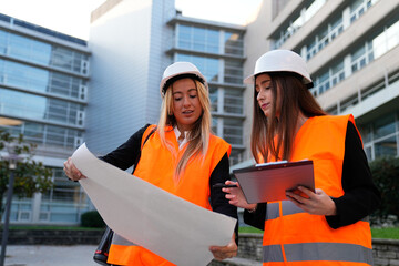 Two women construction engineers wearing hard hats and safety vests are collaborating on building plans outdoors