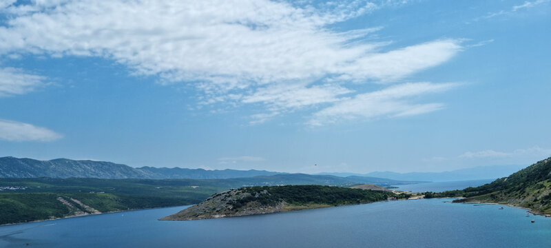 A panoramic view from the Krk Bridge, showcasing the serene Voz Bay on Krk Island with its calm blue waters nestled against a rugged, green coastline under a bright sky