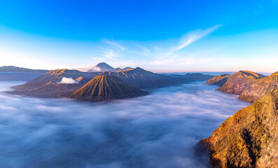 Panoramic early morning view of Mount Bromo in East Java, Indonesia. Smoke curls up from one of the active volcanoes. Fog covers the surrounding desert valley. Blue sky above. 
