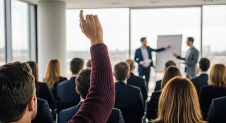 Fototapeta premium An audience member raises their hand during a business presentation, set against a backdrop of large windows and a blurred cityscape view.
