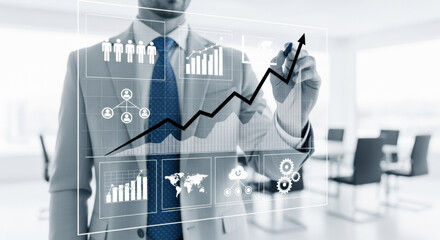 A business man draws a rising graph on a transparent screen in a bright office. The image focuses on the concept of business growth, future and strategy.
