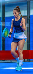 A female padel player walks across a blue indoor court, lamenting a bad play during a competitive match.