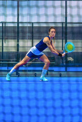 A determined and focused woman, dressed in sportswear, stretches to prepare to receive a ball during a match on a blue indoor court.
