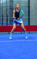A young, athletic woman, dressed in padel attire, stands ready, holding her racket on a blue indoor court.