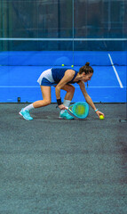 A young female padel player wearing a sports skirt bends down smiling to pick up a ball on the court.
