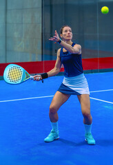 A determined young female padel player prepares herself in an excellent position for a powerful shot on an incoming ball on an indoor blue court.
