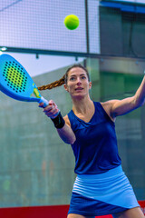 Close-up of a determined and confident female padel player just before returning the ball with a powerful forehand on a blue indoor court.